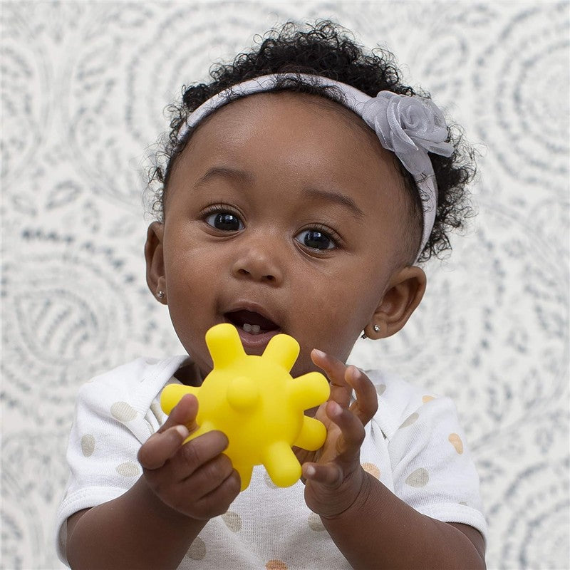 A baby holds a yellow, textured toy resembling a spiky ball, smiling and looking at the camera, with a patterned background.