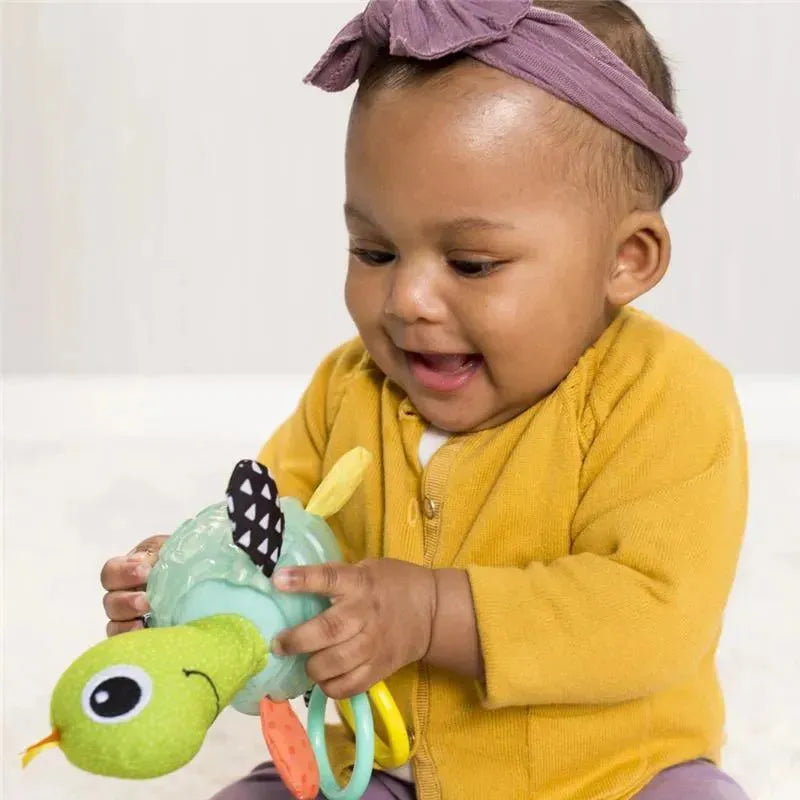 Infantino Textured Sensory Pal Turtle toy held by a cheerful baby wearing a yellow cardigan and purple headband.