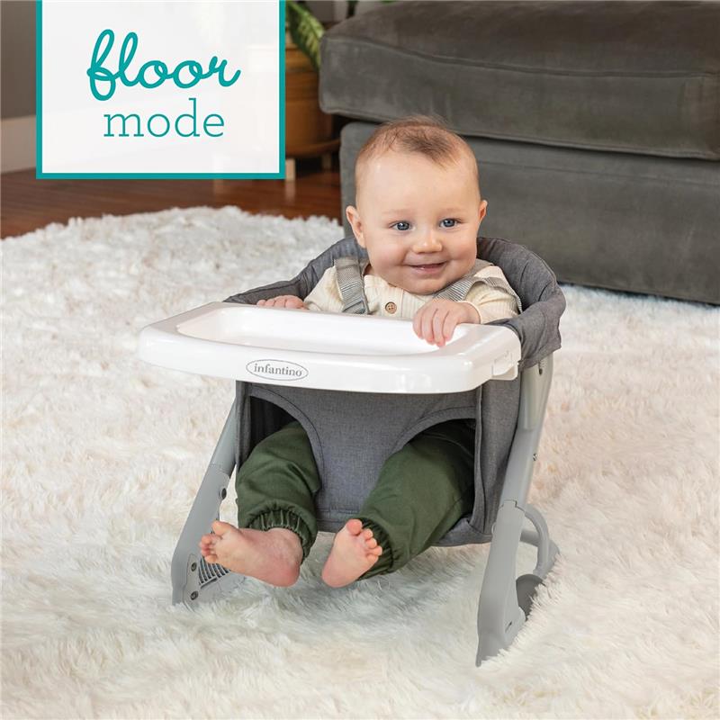 A smiling baby seated in the Infantino Ultimate Travel Booster high chair in floor mode, with a white tray and gray fabric.