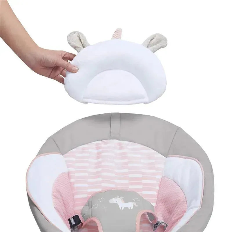 A hand is holding a soft, white unicorn head support for the Ingenuity Soothing Baby Bouncer, showcasing its gentle design.
