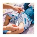 Light blue swim trunks with dinosaur patterns are being put on a child lying on a beach towel.