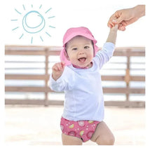 Infant wearing a pink sun hat and pink dragon fruit swim diaper, smiling while being held by an adult outdoors.
