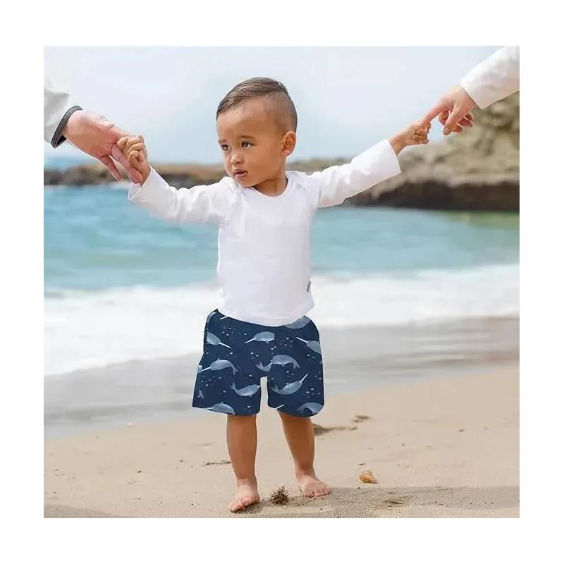 Toddler wearing navy swim trunks with narwhal print and a white long-sleeve shirt, standing on the beach holding hands with adults.