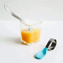 IPlay Learning Spoon Set in aqua next to a glass containing orange puree, showcasing a stainless steel spoon and silicone handle.