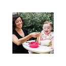 A woman feeds a smiling baby in a high chair using a colorful spoon and a pink bowl, both part of the IPlay Learning Spoon Set.