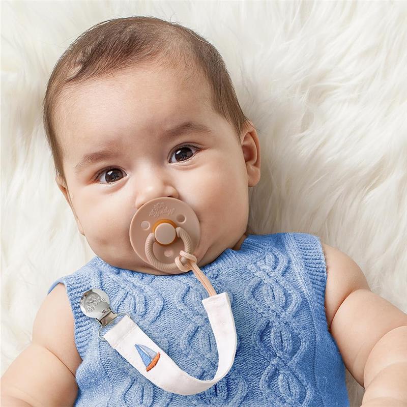 Baby with brown eyes and light brown hair wearing a blue knitted top, holding a beige pacifier attached to a striped fabric strap.