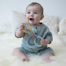 Baby sitting on a soft surface, smiling while holding a set of colorful silicone linking rings in various pastel colors.