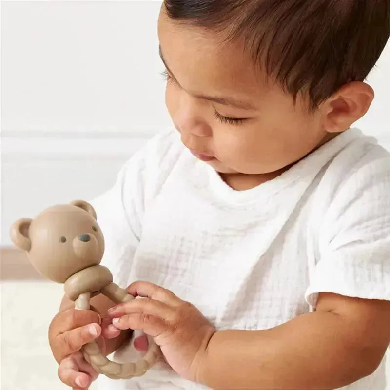 A baby wearing a white shirt holds a beige bear-shaped rattle, examining it closely.