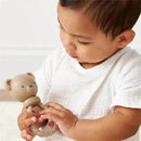 A baby wearing a white shirt holds a beige bear-shaped rattle, examining it closely.