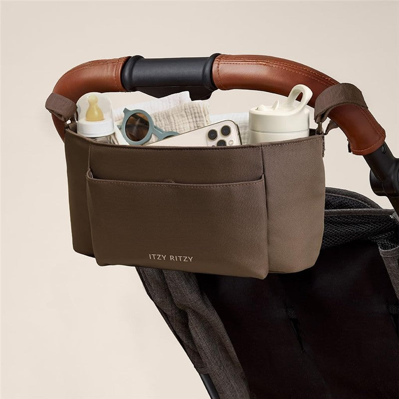 Itzy Ritzy Belong Carry-All Caddy attached to a stroller, holding bottles, sunglasses, and a smartphone, with a brown fabric exterior.