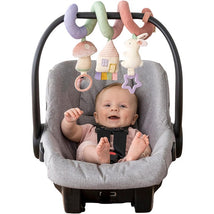 Infant in a car seat smiles, playing with colorful hanging toys including a mushroom, house, and bunny.