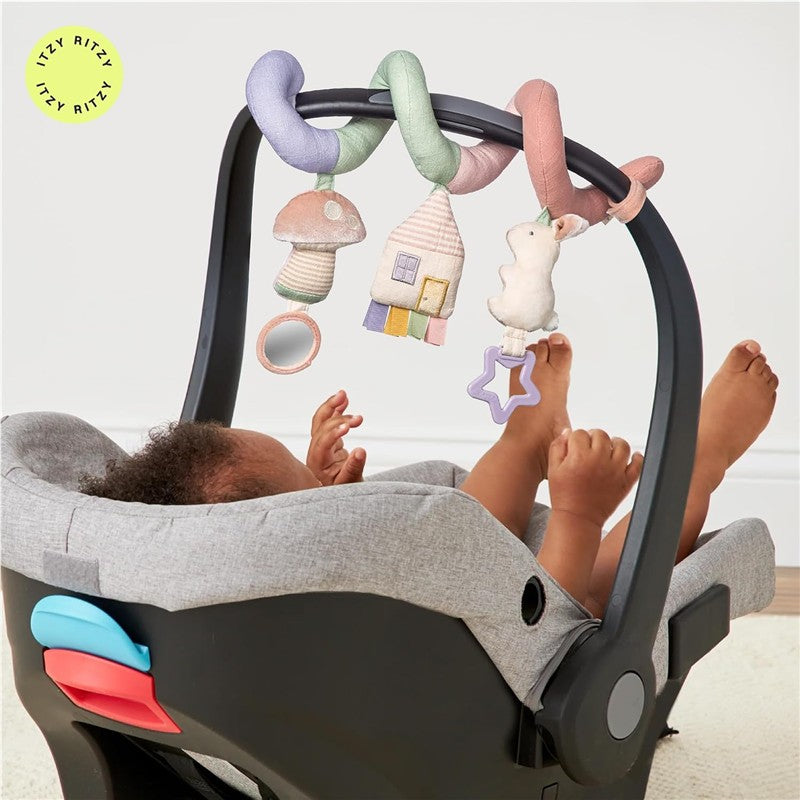 Pastel-colored Itzy Ritzy Bitzy Bespoke Itzy Bitzy Spiral with assorted hanging toys, attached to a baby carrier with a child reaching for them.
