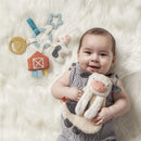 Baby smiling while holding a plush cow toy, surrounded by colorful farm-themed teethers and a barn toy on a fluffy white surface.