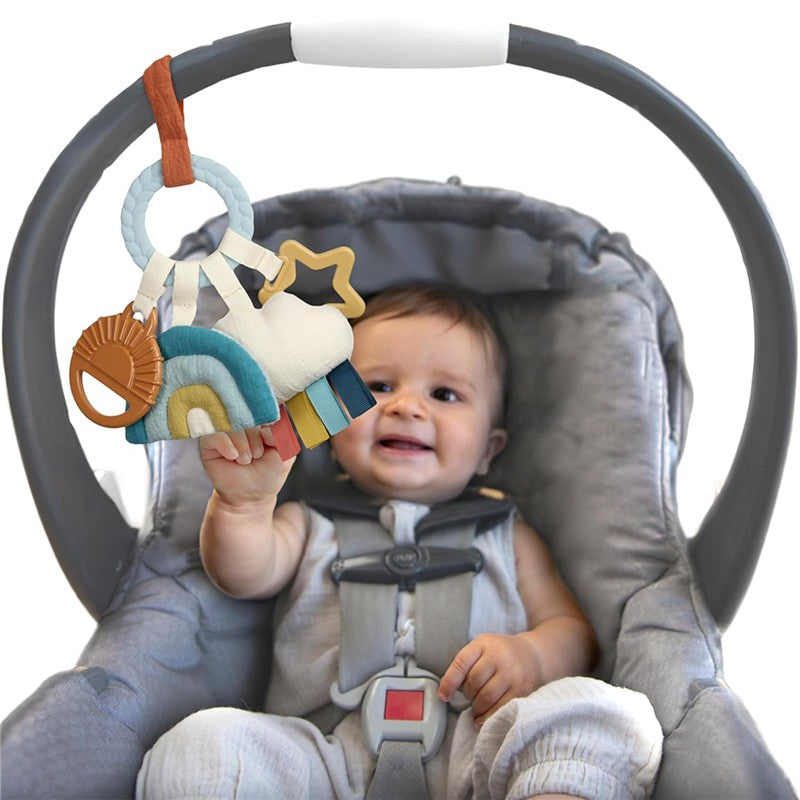 Bitzy Busy Ring teething toy featuring a cloud design, rainbow colors, and various textures, held by a smiling baby in a car seat.