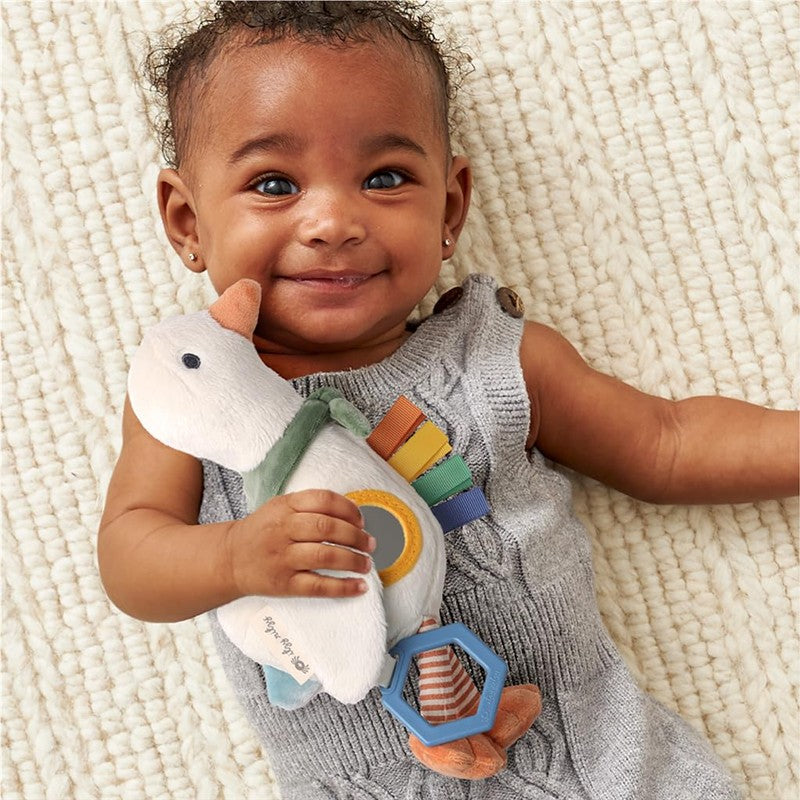 Baby smiling while holding a soft, colorful duck toy with a teething ring, wearing a gray sleeveless outfit.