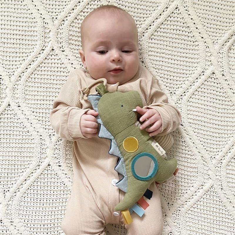 Baby lying on a textured blanket, holding a green dinosaur plush toy with various colored tags and a mirror.