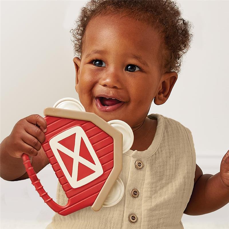Smiling baby holding a red and beige rattle shaped like a barn, featured in a playful, light-filled setting.