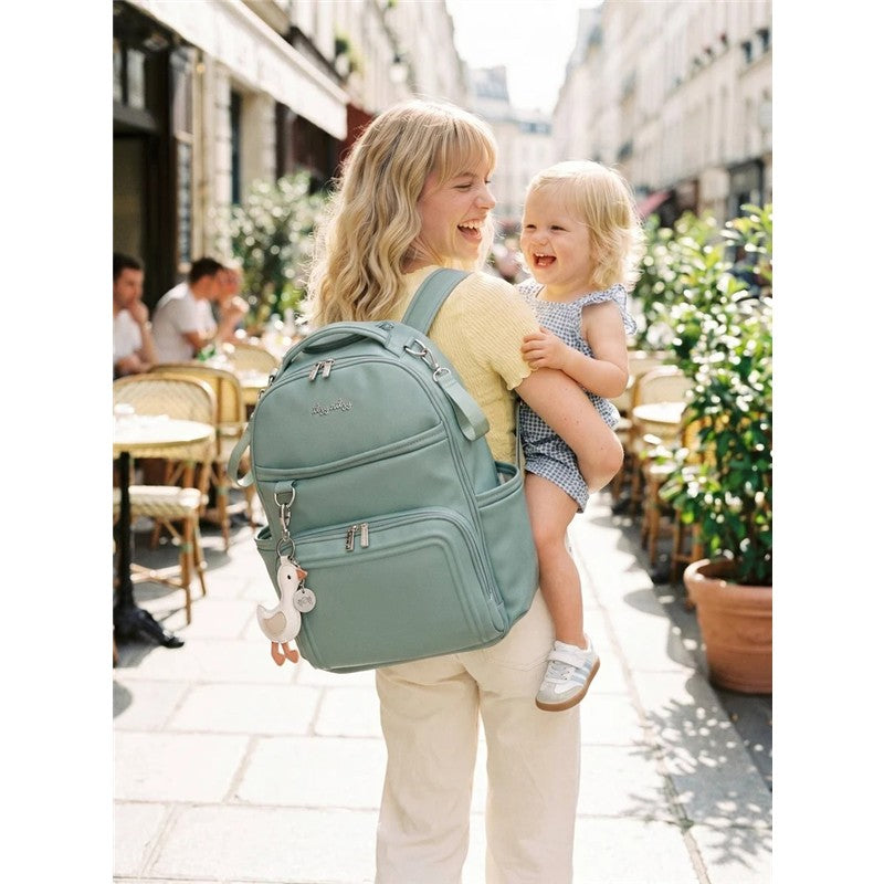 A woman with long hair smiles while carrying a toddler on her hip, wearing a light green Itzy Ritzy Boss Plus™ Backpack Diaper Bag.