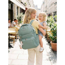 A woman with long hair smiles while carrying a toddler on her hip, wearing a light green Itzy Ritzy Boss Plus™ Backpack Diaper Bag.