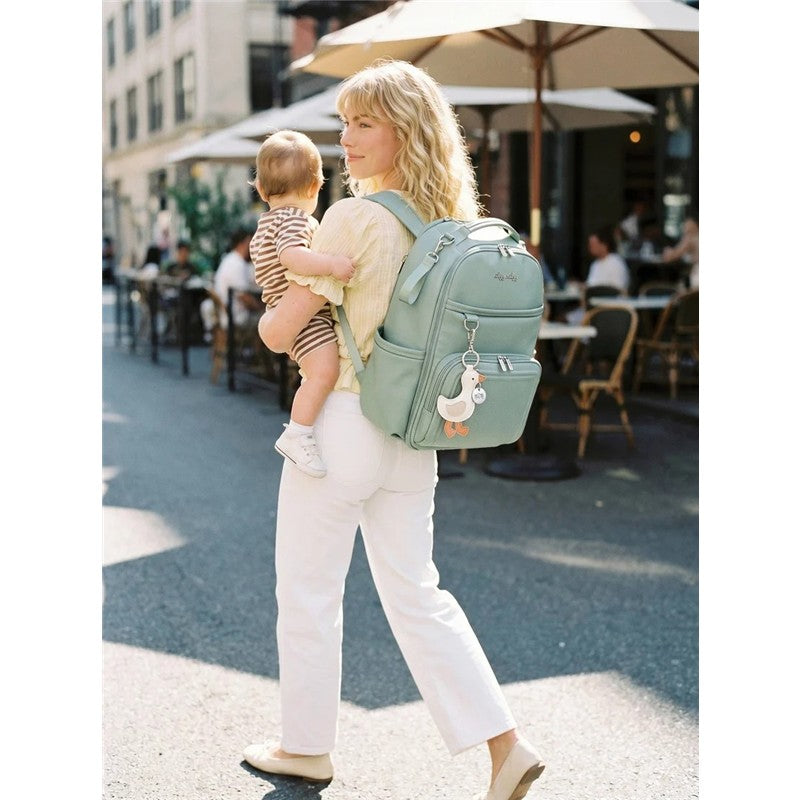 Woman carrying a baby in striped clothing while wearing a mint green Itzy Ritzy Boss Plus™ Backpack Diaper Bag with a duck charm.