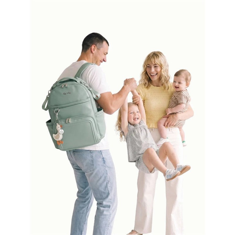 A family interacts joyfully outdoors with a man holding a green Itzy Ritzy Boss Plus™ Backpack Diaper Bag, featuring a cute duck keychain.