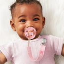 A baby with curly hair smiles while holding a pink pacifier attached to a bow-patterned fabric clip.