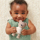Smiling baby holds a beige bear-shaped teether while wearing a green sweater, against a textured white background.