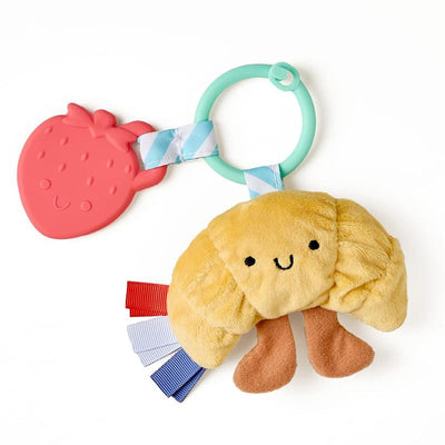 Infant toy and teether featuring a plush croissant with smiling face, attached to a strawberry-shaped teether and colorful ribbons.