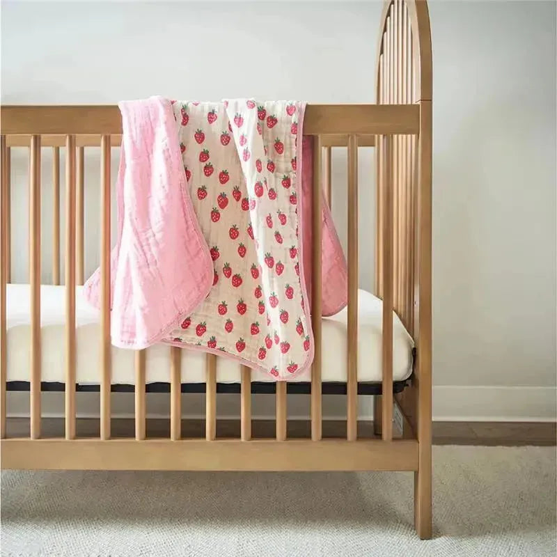 Itzy Ritzy Itzy Blanket™ in Strawberries + Cream draped over a wooden crib, featuring a pink border and strawberry print.