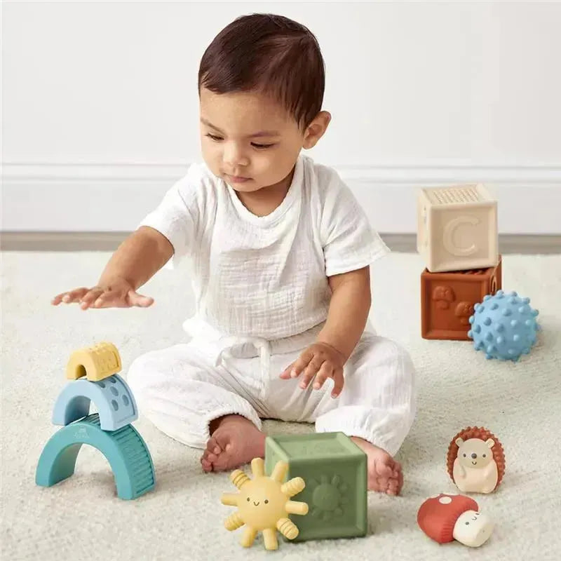 A baby plays with Itzy Ritzy Itzy Blocks™, which include colorful stacking blocks and toy figures on a soft surface.