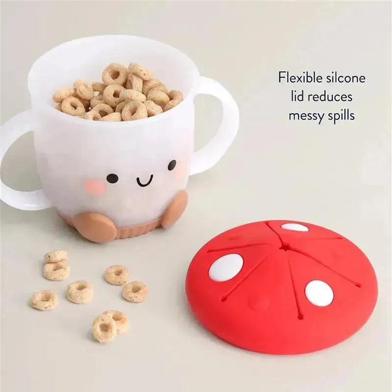 Itzy Ritzy Snack Cup™ in mushroom design, featuring a silicone lid and filled with cereal, with scattered pieces nearby.