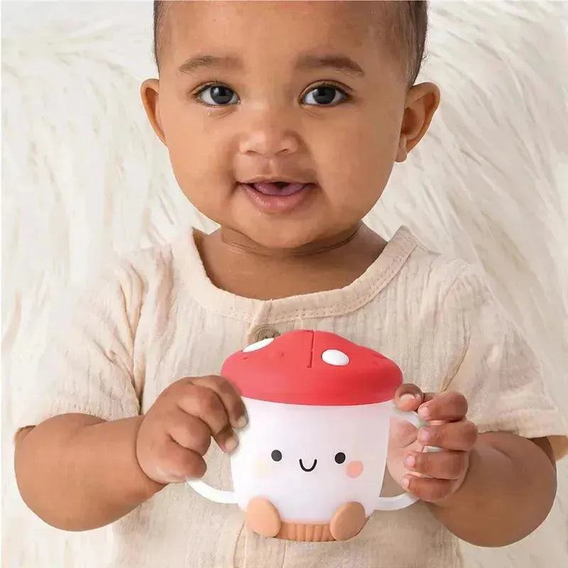 Toddler smiling while holding the Itzy Ritzy Itzy Snack Cup™, designed like a mushroom with a red cap and smiling face.