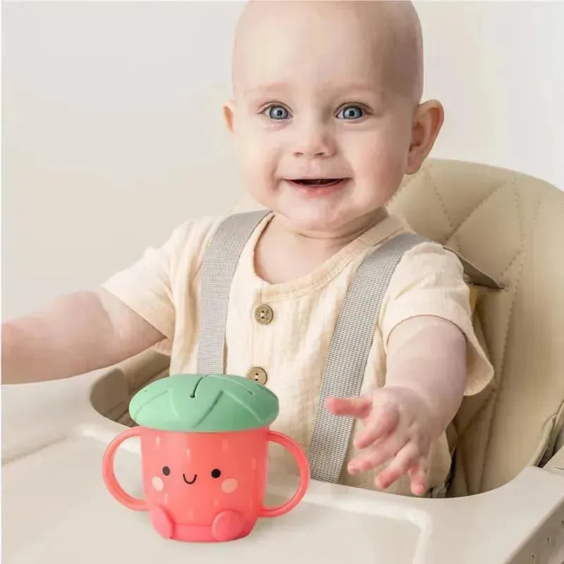 Smiling baby reaching for a pink Itzy Ritzy Snack Cup™ shaped like a strawberry with a green lid, sitting in a high chair.