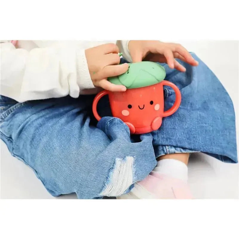 Toddler wearing a white sweater holds an Itzy Ritzy Snack Cup™ in strawberry design with a green lid and smiling face.