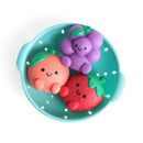 Colorful silicone bath toys shaped like a strawberry, orange, and grape, arranged in a teal colander.