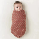 A baby wrapped in a brown patterned multi-use cover, appearing content with a curious expression.