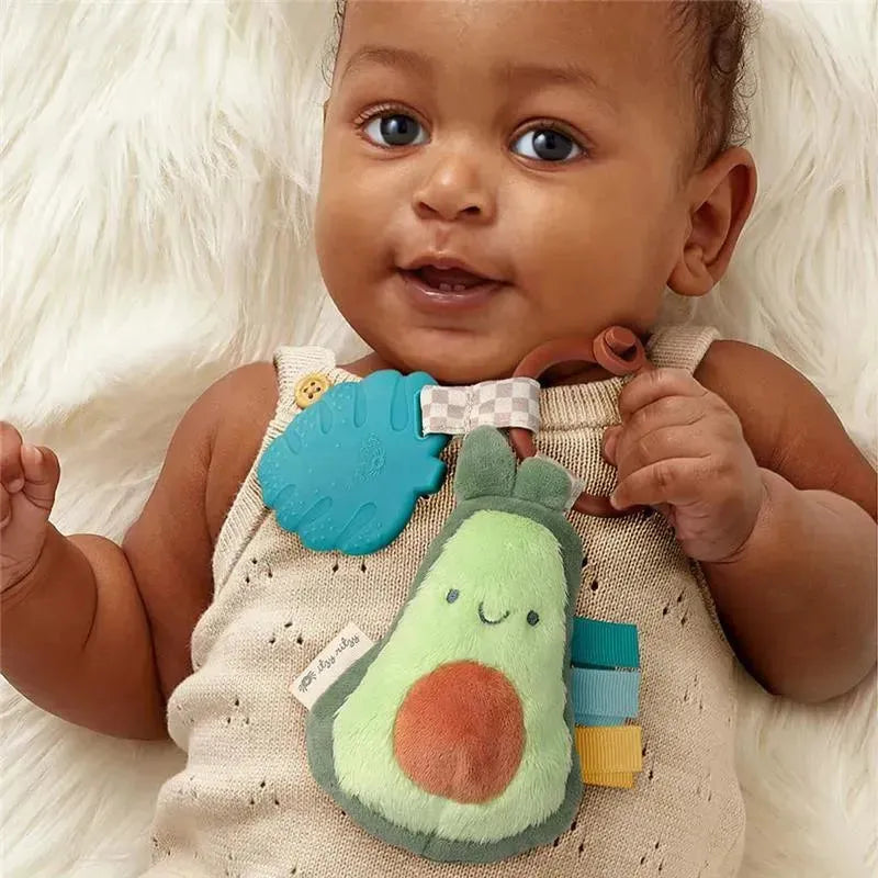 Baby smiling while holding an Itzy Ritzy Pal™ Plush + Teether in avocado shape, featuring soft textures and colorful tags.