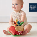 Infant seated on a soft surface, smiling while holding colorful sensory scarves emerging from a red barn-shaped toy.