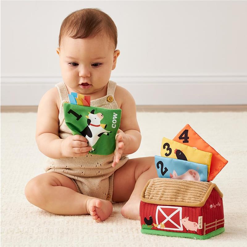 A baby holds a colorful fabric card featuring a cow while exploring a plush barn with numbered animal cards.