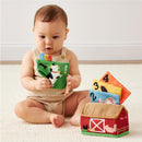 A baby holds a colorful fabric card featuring a cow while exploring a plush barn with numbered animal cards.