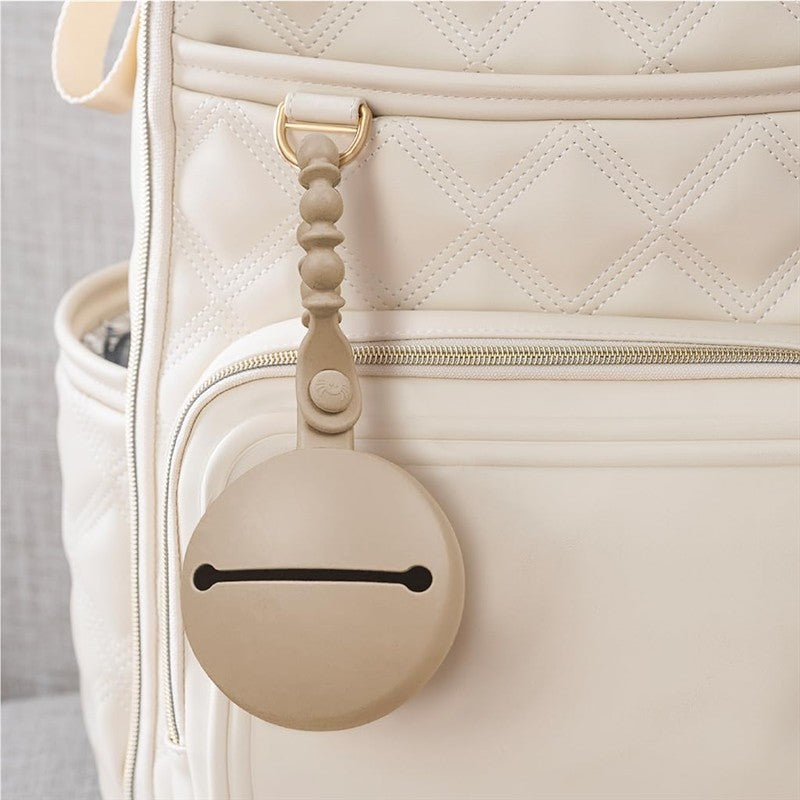 Itzy Ritzy Paci Pod Silicone Pacifier Case in Toast color attached to a cream diaper bag, showcasing its round design and strap.