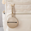 Itzy Ritzy Paci Pod Silicone Pacifier Case in Toast color attached to a cream diaper bag, showcasing its round design and strap.