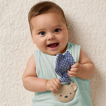 Baby smiling while holding Itzy Ritzy Silicone Teether with Crinkle Tie in bear shape, featuring a blue gingham tie.