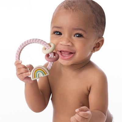 Baby smiling while holding a colorful Itzy Ritzy silicone teether with a rattle, featuring a rainbow design.
