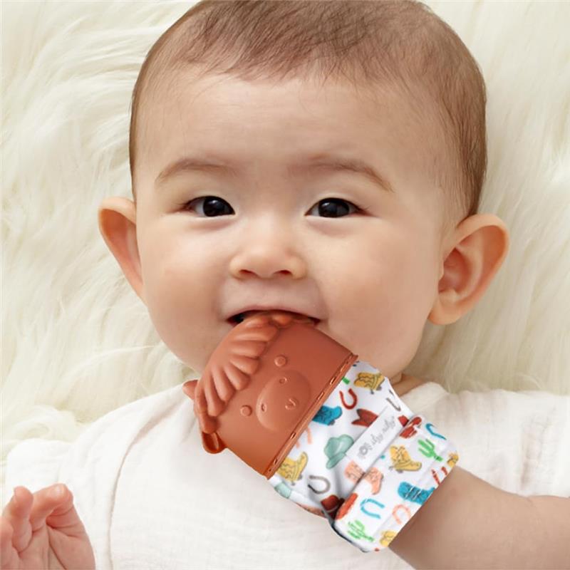 Baby smiling while holding a cow-themed silicone teething mitten, featuring colorful patterns on the fabric.