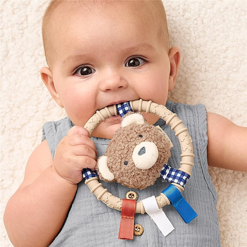 Baby holding an Itzy Ritzy silicone teething ring featuring a plush bear character, with colorful ribbons attached.