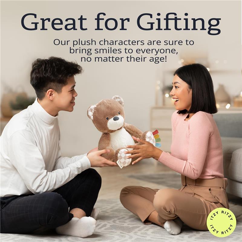 A young man and woman sit on a rug exchanging an Itzy Ritzy Snuggles Plush Toy bear, smiling joyfully.