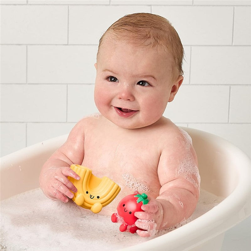 Baby in a bath holding yellow pasta and red tomato squirter toys from the Itzy Ritzy Splash Pals™ set, surrounded by bubbles.