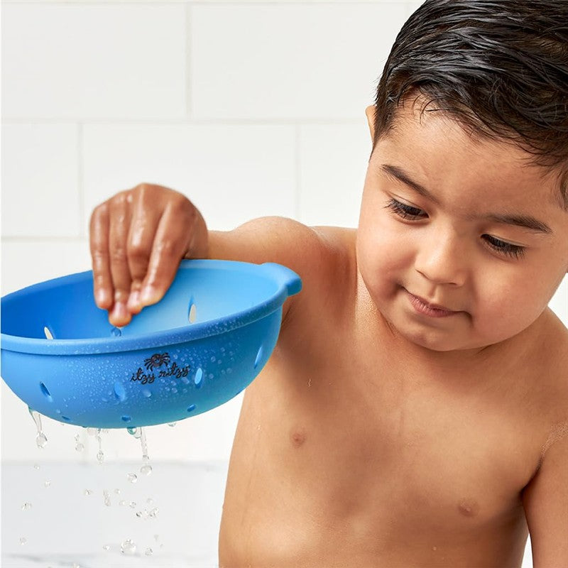 Child playing with a blue Itzy Ritzy Splash Pals™ colander in the bathtub, water dripping from it.