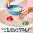 Hands holding a blue strainer filled with colorful, soft bath toys, including a bear and a tomato, in a bubbly bathtub setting.